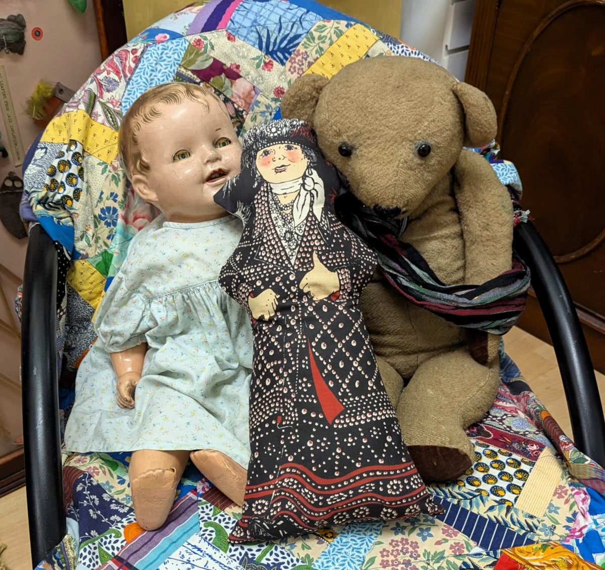 Pearly queen stuffed doll sat between an old toy doll and an old teddy bear, on a chair covered with a patchwork quilt.
